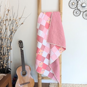 May include: A pink and white patchwork quilt draped over a bamboo ladder. The quilt features a variety of pink shades and white squares. A guitar leans against the wall, and decorative branches are in a vase.