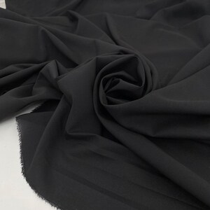 Peut inclure: Gros plan sur un tissu noir, drapé et plié. Le tissu présente une texture lisse et un léger brillant, avec des plis et des ombres qui créent de la profondeur. Les bords du tissu sont bruts et non finis, ajoutant une touche de texture.