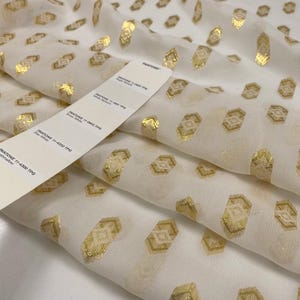 Op de afbeelding: Witte stof met een herhalend gouden geometrisch patroon. Een Pantone kleurstalenkaart ligt op de stof en toont kleurnamen als "Snow White" en "Marshmallow". De stof lijkt licht en vloeiend.