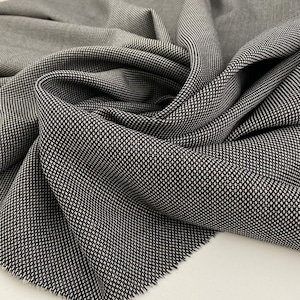 Op de afbeelding: Close-up van een textiel met een zwart-wit stippenpatroon. De stof is gedrapeerd en toont plooien en variaties in licht en schaduw. Het materiaal lijkt van een gemiddeld gewicht te zijn, geschikt voor kleding of woondecoratieprojecten.