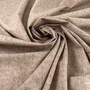 Op de afbeelding: Beige stof met een herhaald patroon van bruine bladeren. De stof is gevouwen en gedrapeerd, waardoor schaduwen en textuur ontstaan. De bladeren zijn gestileerd en gerangschikt in een natuurlijk, organisch ontwerp. Ideaal voor naai- en knutselprojecten.