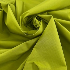 May include: Close-up of a vibrant green fabric with a subtle grid pattern. The material is tightly woven, with visible folds and a swirling arrangement, creating a textured and dynamic visual effect. The fabric has a slight sheen.