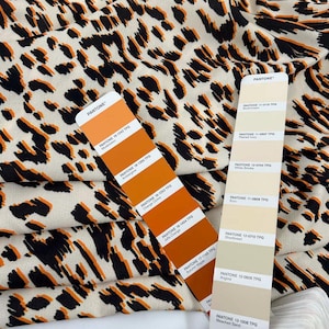 May include: Cream-coloured fabric with a black and orange animal print pattern. Two Pantone colour charts are visible, displaying shades of orange and cream. The fabric is suitable for clothing or home furnishings.