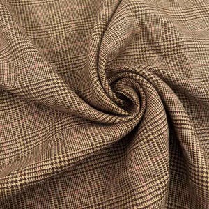 May include: A woven fabric featuring a classic brown and tan glen plaid pattern with subtle, thin pink accent lines running through the check design. The material is gathered in a soft, draped fold, highlighting the intricate, repeating grid texture.
