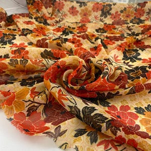 May include: A flowing fabric with a floral pattern in shades of red, orange, brown, and black on a cream background. The fabric is draped and folded, showcasing the intricate design of the flowers and leaves. Ideal for apparel or home decor.