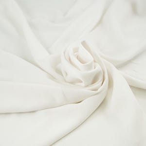 以下が含まれることがあります： 柔らかい折り目と中央にバラのような配置が施された白い生地のクローズアップ。生地は滑らかな質感とマットな仕上がりで、繊細でエレガントな外観を作り出しています。全体的な効果は、穏やかな動きと微妙な光の遊びです。