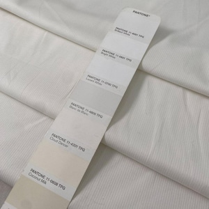 May include: Close-up of white fabric with a ribbed texture. A Pantone color swatch is placed on top, displaying shades of white, including "Brilliant White," "Bright White," and "Coconut Milk." The fabric appears soft and slightly wrinkled.