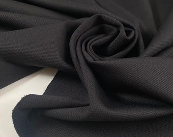 May include: Close-up of a dark charcoal-colored fabric with a textured, woven pattern. The fabric is draped and folded, creating a swirling effect. The material appears to be a medium-weight textile, suitable for apparel or home decor.