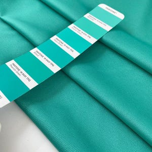 Puede incluir: Primer plano de tela turquesa doblada con una carta de colores Pantone. La carta muestra varios tonos de turquesa, incluyendo Electric Green, Aqua Green, Billiard y Peacock Green. La tela tiene una sutil textura acanalada.