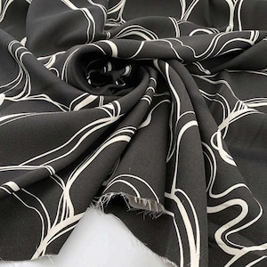 May include: Black fabric featuring a white, abstract, swirling design. The material is draped and folded, highlighting the texture and pattern. The edges of the fabric are visible, suggesting it could be a cut piece or a roll of fabric.