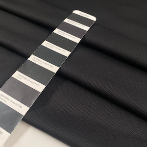 May include: Close-up of black fabric with a textured weave. A Pantone color chart is placed diagonally across the fabric, displaying shades of gray and black. The chart includes color names like "Black Onyx" and "Caviar".
