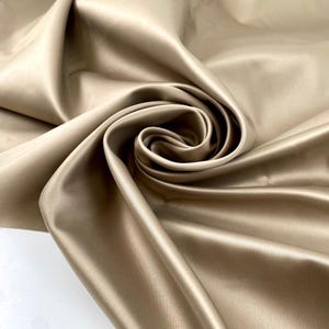 May include: Close-up of a swirl of beige satin fabric. The fabric is smooth and reflective, with soft folds and shadows creating depth. The color is a warm, neutral tone, suitable for various sewing and crafting projects.