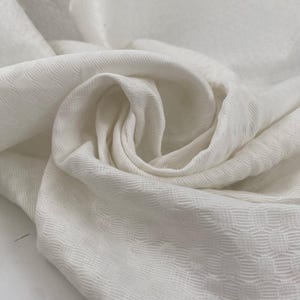 Puede incluir: Primer plano de una tela blanca con un patrón texturizado y tejido. La tela está drapeada en pliegues suaves, creando sombras y reflejos. El material parece ligero y fluido, con un brillo sutil.