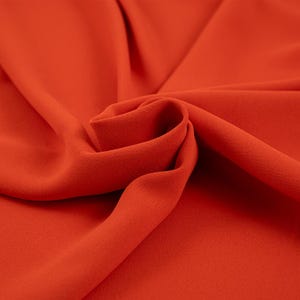 Op de afbeelding: Close-up van een gladde, feloranje stof. De stof is gevouwen en gedrapeerd, waardoor de textuur en kleur zichtbaar zijn.
