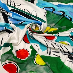Op de afbeelding: Witte stof met een abstract ontwerp. Het heeft gedurfde streken in blauw, groen, geel en rood, met zwarte lijnen en vormen. De stof is gevouwen, waardoor schaduwen en textuur ontstaan, ideaal voor diverse textielprojecten.
