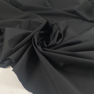 May include: A close-up of a black fabric with a soft, matte finish. The fabric is draped and folded, creating shadows and texture. The edges of the fabric are visible, showing the material's weave. The overall appearance suggests a textile suitable for clothing or home decor.