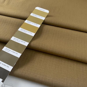 Puede incluir: Primer plano de tela beige doblada con una carta de colores Pantone. La carta de colores muestra tonos de oro, bronce y beige, incluyendo "Dull Gold" y "Antique Bronze". La tela tiene un sutil patrón de cuadrícula.