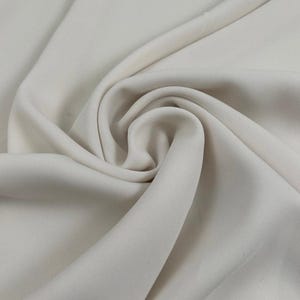 Puede incluir: Una tela de color blanco roto sólido con una textura suave y mate, dispuesta en una serie de pliegues y drapeados suaves que crean sombras y reflejos naturales en la superficie, enfatizando el color consistente y el movimiento del material.