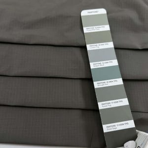 Op de afbeelding: Close-up van een textiel, donkergrijze stof met een rasterpatroon. Een Pantone-kleurenkaart toont verschillende tinten grijs en groen, waaronder "London Fog" en "Dried Sage."