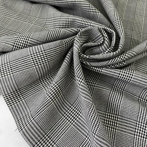 Può includere: Primo piano di un tessuto a quadri bianco e nero. Il tessuto presenta un classico motivo a quadri scozzesi, con una trama fine. Il tessuto è piegato, rivelando la sua consistenza e l'intricato motivo. Il tessuto sembra essere di peso medio.