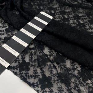 May include: A close-up shot of black lace fabric with a floral design. The fabric is arranged in folds, highlighting its texture. A Pantone colour card is included, offering a colour reference for the black material.