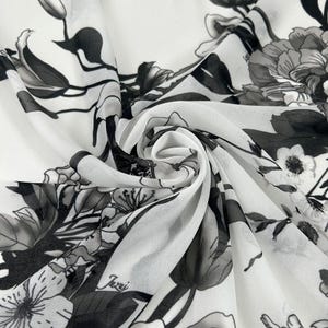 Puede incluir: Tela blanca con un estampado floral en negro y gris. El diseño presenta grandes flores y hojas, creando un contraste llamativo sobre el fondo blanco. La tela está doblada, mostrando la textura y la caída del material. La palabra "Jazui" está impresa en la tela.