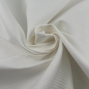 Peut inclure: Un tissu blanc uni présentant un motif subtil de grille en forme de losanges répétitifs. Le matériau est drapé en plis souples et fluides, mettant en valeur la surface texturée et le tissage géométrique constant sur tout le textile.