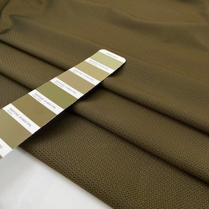 May include: Olive green fabric with a subtle geometric pattern. A Pantone color swatch card displays shades of green, from light to dark. The fabric is folded, showing texture and depth.