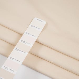 Può includere: Primo piano di tessuto color crema piegato con una cartella colori Pantone. La cartella mostra varie tonalità di bianco sporco e crema, tra cui Whisper White ed Eggnog. Il tessuto ha una trama sottile.