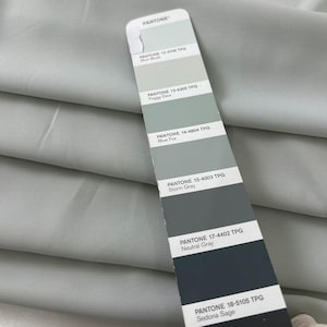 May include: A fabric swatch featuring a palette of grey and blue-green shades, such as "Blue Blush," "Foggy Dew," and "Storm Gray." The Pantone colour guide is shown against a background of folded fabric.