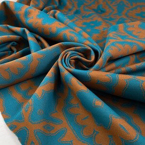 Op de afbeelding: Turquoise stof met een bruin patroon. De stof is gevouwen en laat het patroon in detail zien.