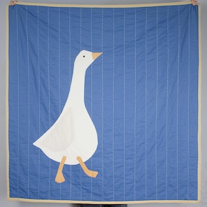 May include: A blue quilted throw blanket with a white goose applique. The goose is walking to the right and has a yellow beak.