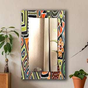 Peut inclure: Un miroir mural rectangulaire doté d'un large cadre décoratif orné d'un motif abstrait coloré composé de formes géométriques, de lignes et de motifs floraux dans des tons orange, vert, noir, blanc et bleu sur un fond sombre.