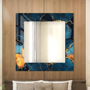 Pode incluir: Um espelho quadrado com uma moldura de design abstrato azul e dourado. O espelho está pendurado em uma parede de madeira acima de uma cama com almofadas marrons.