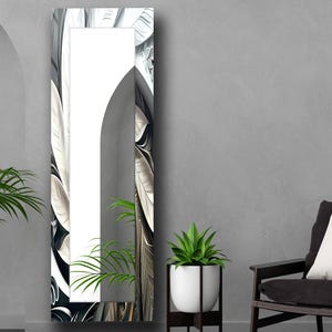 Pode incluir: Um espelho alto e retangular com uma parte superior arqueada. A moldura apresenta um design floral em preto e branco. O espelho é exibido contra uma parede cinza, com uma cadeira e uma planta em vaso em primeiro plano.