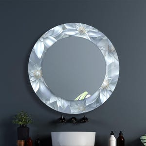 Peut inclure: Un miroir rond avec une bordure à motif floral. La bordure présente des fleurs blanches avec des accents gris et argentés. Le miroir est monté au-dessus d'un lavabo blanc et d'une étagère en bois dans une salle de bain.