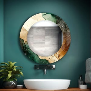 Op de afbeelding: Ronde decoratieve spiegel met een aquarelontwerp in groen, goud en bruin. De spiegel is gemonteerd op een teal muur boven een witte wastafel en houten aanrecht. Een potplant en accessoires zijn ook zichtbaar.