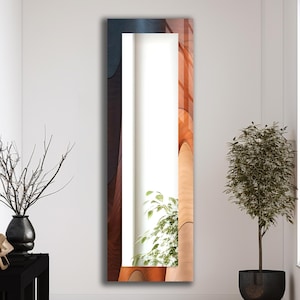 May include: A tall, rectangular mirror with a decorative frame. The frame features a layered design with shades of brown, orange, and blue. The mirror is positioned against a white wall, reflecting a glimpse of a plant.