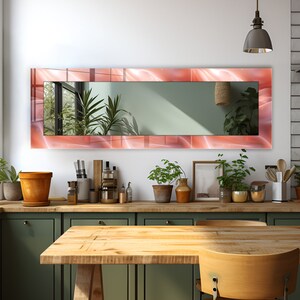 Puede incluir: Un espejo rectangular largo con un marco de diseño abstracto rosa y blanco. El espejo está colgado en una pared blanca sobre una encimera verde con una mesa de madera y sillas delante.