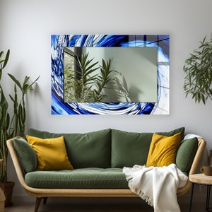 Puede incluir: Un espejo rectangular con un diseño abstracto de espiral azul y blanco. El espejo está colgado en una pared blanca sobre un sofá verde con almohadas amarillas.