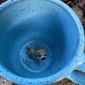 Könnte beinhalten: Eine blau gesprenkelte Keramik-Tasse mit einer kleinen grauen Hundefigur im Inneren. Die Tasse hat einen Henkel und steht vor einem natürlichen Hintergrund. Innen und außen ist die Tasse blau mit dunkelblauen Sprenkeln.