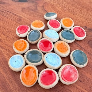 Könnte beinhalten: Eine Sammlung kleiner Keramiksteine mit gesprenkelter, cremefarbener Basis und glänzendem, farbigem Zentrum. Die Farben umfassen Rot, Orange, Blau und Türkis. Die Steine sind auf einer dunklen Holzoberfläche angeordnet.