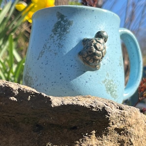 Könnte beinhalten: Eine hellblaue Keramik-Tasse mit einer angebrachten Schildkrötenfigur. Die Tasse hat eine gesprenkelte Textur und einen gebogenen Henkel. Die Schildkröte ist braun und hat einen strukturierten Panzer. Die Tasse steht auf einem Felsen.