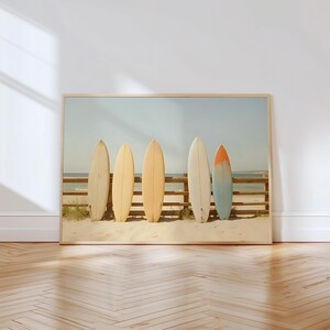 Könnte beinhalten: Ein gerahmter Druck mit fünf Surfbrettern, die an einem Holzzaun an einem Sandstrand lehnen. Die Surfbretter sind in verschiedenen Farben gehalten, darunter Creme, Gelb und Orange. Der Himmel ist hellblau, was eine sommerliche Atmosphäre erzeugt.
