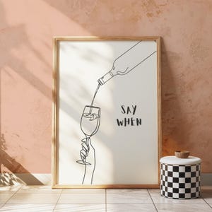 May include: A framed art print with a minimalist line drawing of wine being poured into a glass. The words "SAY WHEN" are written in a handwritten font. A black and white checkered container with a wooden lid sits nearby.