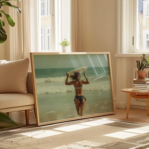 May include: A framed print of a person in a bikini carrying a surfboard into the ocean. The image is in a light wooden frame, leaning against a wall in a well-lit room. The ocean is visible in the background.