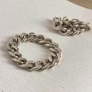 May include: A silver-colored chain ring and a small chain segment. The ring is made of interlocking oval links, creating a chunky, textured appearance. The chain segment is a smaller version of the ring's design. Both pieces are set against a white background.