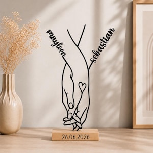 Pode incluir: Uma escultura de arte linear em metal preto que retrata duas mãos dadas com o contorno de um pequeno coração. Os nomes Mayleen e Sebastian estão integrados no design. A escultura está montada numa base de madeira retangular com 26.06.2026.