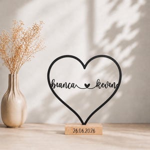 Könnte beinhalten: Ein schwarzes, herzförmiges Schild mit den Namen "bianca" und "kevin", die durch ein kleines Herzsymbol in der Mitte verbunden sind. Das Schild ist auf einem rechteckigen Holzsockel montiert, auf dessen Vorderseite das Datum "26.06.2026" eingraviert ist.