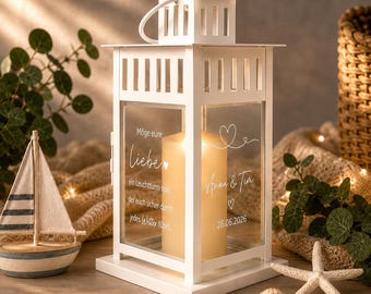 May include: A white lantern with a handle and glass panels, holding two lit candles. The glass displays text and a heart design. A small sailboat, starfish, and seashell are in the foreground. The lantern is on a wooden surface.
